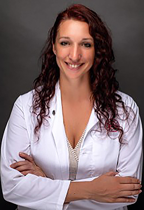 Dr. Melissa Ballantyne smiles with her arms crossed in front of her. She is standing in front of a dark studio background and is wearing a tan top and white lab coat.