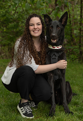 Dr. Madeline Hughley smiles outside in a wooded area. She is on one knee, hugging her German shepherd. She is wearing black jeans and a grey cardigan.