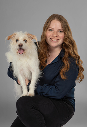 Dr. Sierra LeFore smiles for a studio portrait while kneeling and holding her white terrier mix. She is wearing black dress pants and a navy blouse.
