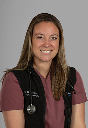 Dr. Shannon Miller smiles for a professional headshot in front of a grey studio background. She is wearing heather maroon scrubs, a black vest and has a stethoscope around her neck.
