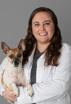 Dr. Tori Rastl smiles for a professional photo while holding her tan and white Rat Terrier mix in her arms. She's wearing dark grey scrubs and her BluePearl whitecoat.