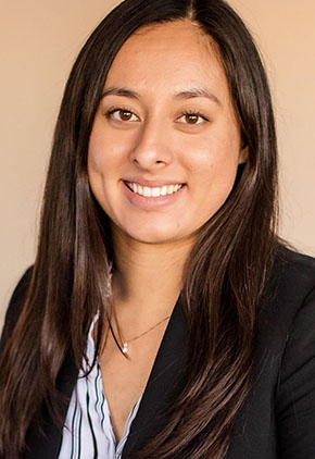 Dr. Alexis Urrea smiles for a professional headshot in front of a tan background. She is wearing a multicolored dress shirt and black blazer.