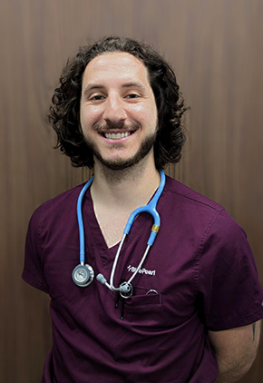 Dr. Alex Ortiz Colosi smiling in front of a dark wood wall. He is wearing maroon scrubs and has a blue stethoscope around his neck.