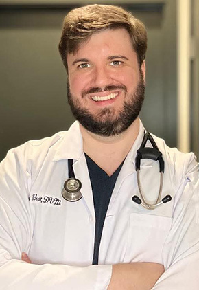 Dr. Steven Butt in a clinic hallway, leaning against the wall with his arms crossed. He's smiling, wearing his white coat and has a stethoscope around his neck.