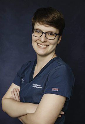 Dr. Kelly Schrock, wearing glasses and teal scrubs with the BluePearl Pet Hospital logo, smiles with arms crossed while posing against a dark blue background.
