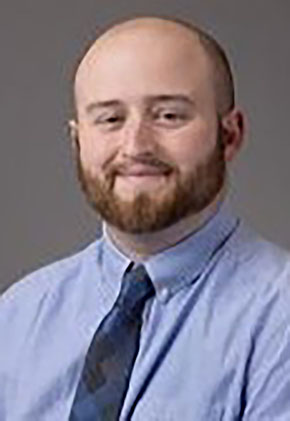 Dr. Andy Arnold smiles for a professional headshot in front of a dark grey background. He's wearing a light blue dress shirt and a navy tie.