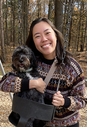 Dr. Leeza Kao smiles outside on a fall day. She is hugging her dog, Rowena, who is sitting in a baby swing seat. Dr. Kao is wearing a patterned knit sweater.