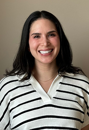 Dr. Sophie Eiger smiling in front of a tan wall. She has on a white sweater with thin black stripes.