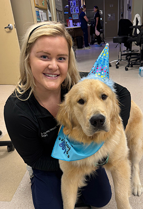 Dr. Courtney Hancock smiles while kneeling next to a golden retriever in the charting station at the hospital. The dog has a happy birthday hat and bandanna on and Dr. Hancock is wearing black scrubs.