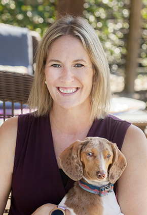 Dr. Julia Spade smiles outside while holding her wiener dog mix. She has on a violet top and there are green trees behind her.