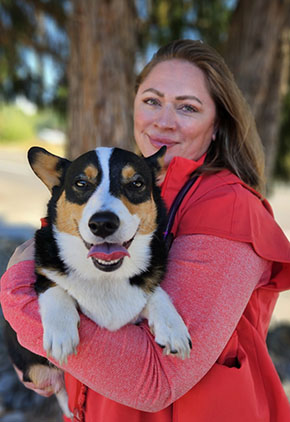 Dr. Shannon Tyler smiles outside in front of a tree. She is wearing red scrubs with a red long sleeve shirt underneath. In her arms is her black, white and brown corgi who looks to be smiling right into the camera.