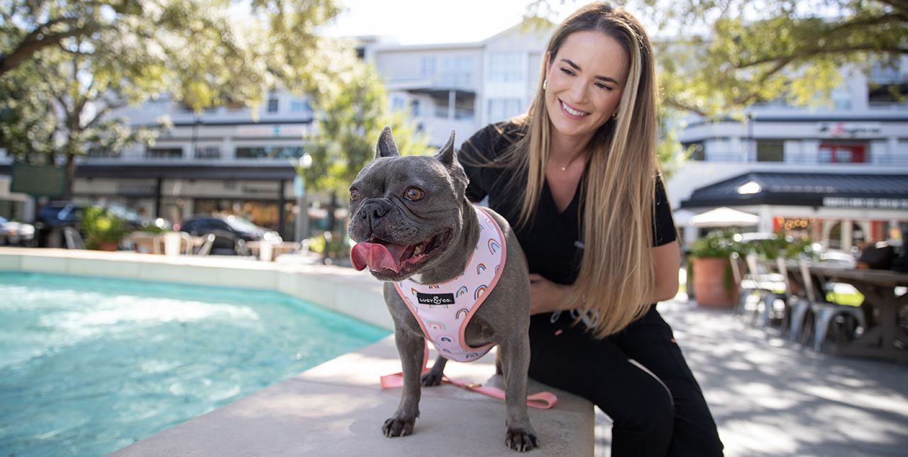 A woman smiles at a French bulldog while they sit on the edge of a fountain in a shopping plaza.