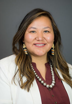Dr. Kaitlyn Benally smiles for a professional headshot in front of a grey background. She is wearing a maroon blouse, beaded necklace and her white coat.