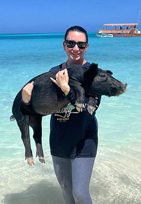 Dr. Lauren Young smiling and standing ankle-deep in the ocean holding a black pig in the Bahamas.