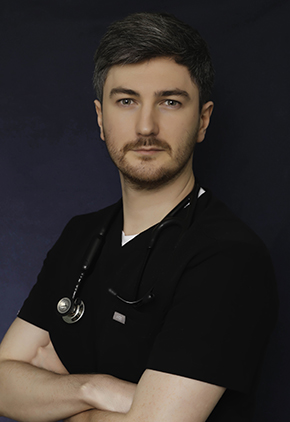 Dr. Jonathan Mullins, with short gray hair and a trimmed beard, wears black scrubs and a stethoscope around his neck while posing against a dark background and crossing his arms.