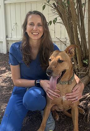 Dr. Sarah Christie kneels outside in her back yard next to her dog. She is wearing blue scrubs.
