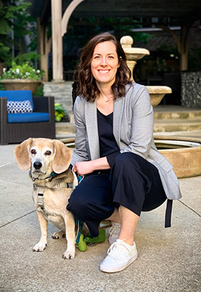 Dr. Barbur outside, in front of a small bubbling water fountain. She is smiling and wearing a grey blazer and black slacks while kneeling next to her beagle, Pixel.