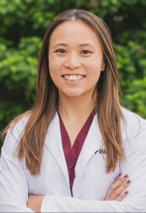Dr. Brittany Ho smiling outside. She has on a maroon top and her BluePearl white coat. She has her arms crossed in front of her.