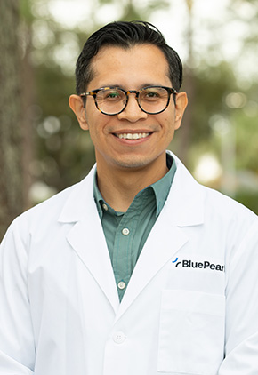 Dr. Miguel Angel Martinez smiling outside. He is wearing a sea green button-up, black glasses and his BluePearl whitecoat.