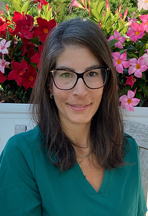Dr. Farrah Horowitz sitting on a bench in front of a tree in bloom with red and pink flowers. She has on teal scrubs, glasses and a smile.