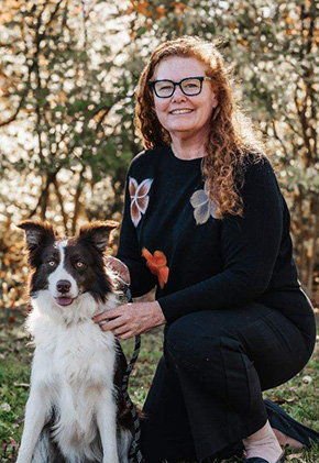 Dr. Jean Rogers kneeling on grass in a sunlit, wooded area, smiling next to her black and white long-haired dog. She is wearing a black sweater with floral designs, black pants and glasses.