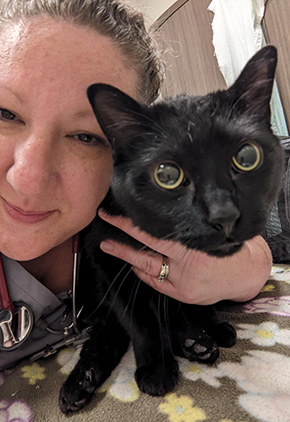 Emily Elmer, CVT, is shown smiling while crouching and holding a black cat close for snuggles.