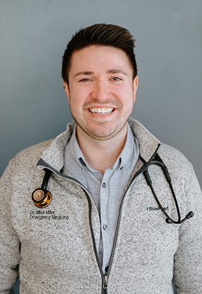 Dr. Mike Miller Smiling in front of a blue-grey wall. He has on a light heather-grey full-zip BluePearl sweatshirt with a light blue button-up and a stethoscope around his neck.