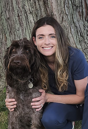 Dr. Madeline Vind smiles outside while kneeling next to her brown shaggy-haired dog.