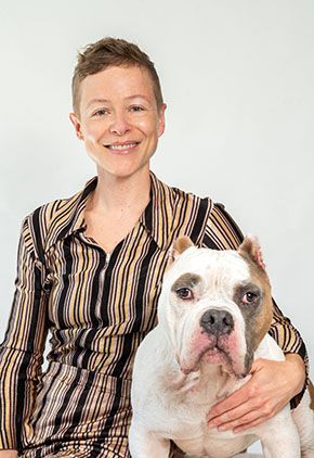 Dr. Emily Farrell smiles for a professional photo sitting next to her bulldog. She is wearing a brown striped dress.