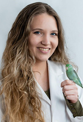 Dr. Kaitlyn Nikirk, wearing a white lab coat, smiles while holding a small green parrot on her hand.