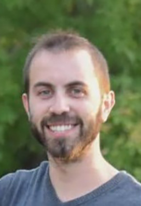 Dr. Aharon Helman smiling outside. There is a green leafy tree in the background and he has on a navy grey shirt.