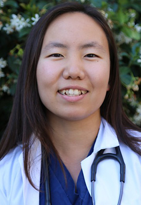 Dr. Adora Min smiling outside in front of a leafy green tree. She is wearing navy scrubs, her white coat and a stethoscope around her neck.