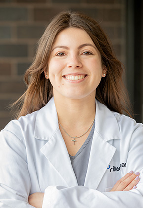 Dr. Michelle Schnabel smiling outside in front of a brick wall. Her arms are crossed and she has on a grey top and her BluePearl white coat.