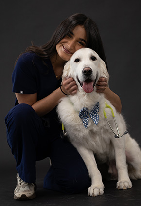 Dr. Claudia Odiot, wearing navy scrubs, kneels beside her golden retriever, Marcelo, in a professional studio photo with a black backdrop. She smiles as she gently holds Marcelo’s head, scratching his ears. Marcelo, wearing a blue bowtie and a stethoscope around his neck, appears to be smiling too.