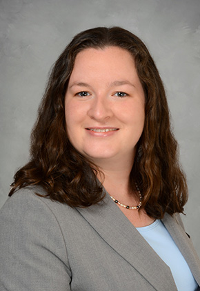 Dr. Kate Goodman smiling in a professional headshot. She is wearing a light blue dress shirt, a black and silver necklace, and a grey blazer, with a standard grey studio backdrop behind her.