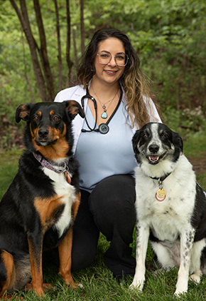 Dr. Elizabeth Bruzinski smiles outside for a professional photo while kneeling between her two dogs. She is wearing black dress pants, a white blouse and has a stethoscope around her neck.