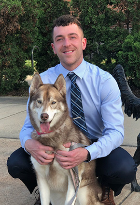 Dr. Ben Kinnamon outside with his Siberian Husky, Nova. He is squatting down in a light blue dress shirt, blue tie, and dark navy dress pants, embracing Nova, who is sitting in front of him with her tongue out.