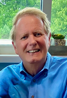 Dr. Thomas Hanson, smiling and wearing a bright blue dress shirt, seated indoors in front of a window with green trees in the background and a small succulent plant on the windowsill.