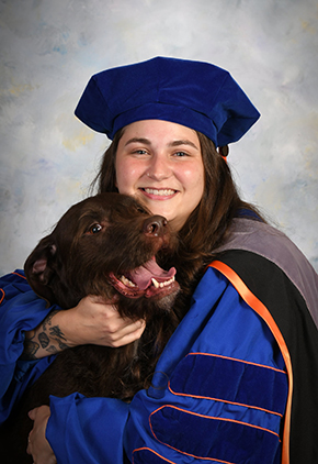 Dr. Crystal Van Horn smiling for a graduation portrait in front of a grey background. She has on her blue and orange doctoral robes, hat and stole. She is hugging her chocolate lab, who has their mouth open in excitement.