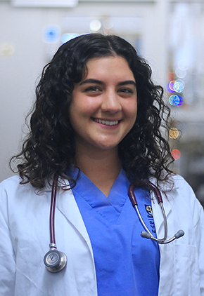 Dr. Nicole Hasan smiling for a photo in the hospital. She has on blue scrubs and her white coat with a stethoscope around her neck.