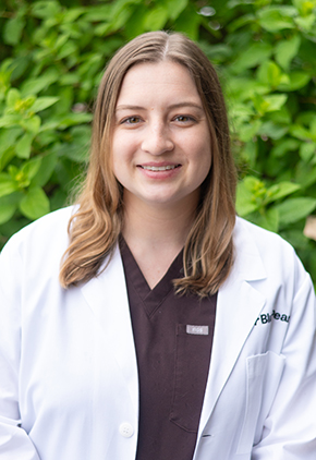 Dr. Milica Waters smiles outside in front of a leafy, green bush. She is wearing maroon scrubs and her BluePearl whitecoat.