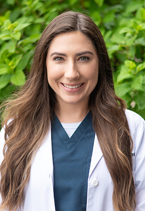 Dr. BreAnna McCartan smiles outside in front of a green, leafy tree. She is wearing dark navy scrubs and her BluePearl whitecoat.