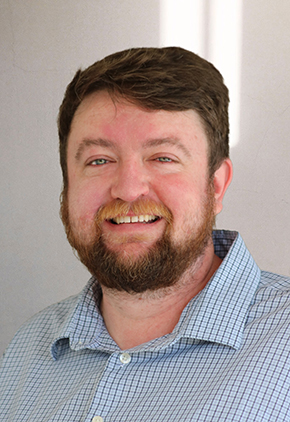 Dr. Bryan Williams smiling for a professional photo in front of a white backdrop. He has on a blue stripped button-up.