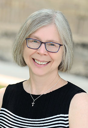 Dr. Kari Anderson-Carlquist smiles outside in front of a building in early spring, with snow visible on the bank behind her. She is wearing a black and white professional sleeveless top and glasses.