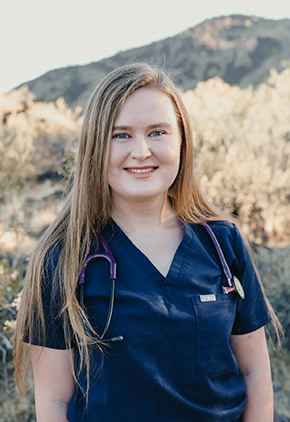 Dr. Caroline Parot smiling outside with a mountain range in the background. She has on navy scrubs with a stethoscope around her neck.