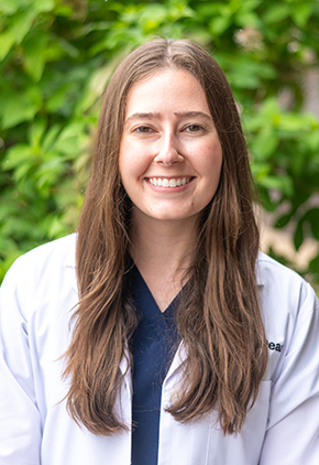 Dr. Hailey Everhart smiles outside in front of a green, leafy tree. She is wearing navy scrubs and her BluePearl whitecoat.