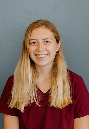 Dr. Maddie McDermott smiling for a professional photo in front of a blue-grey background. She has on a maroon top and a small gold necklace.
