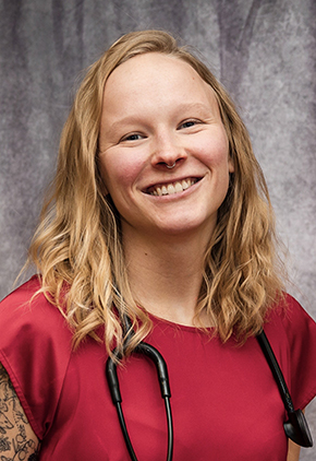 Dr. Dora Uidl smiling in front of a grey background. She has on a red top with her stethoscope around her neck. Tattoos on her right arm are visible.