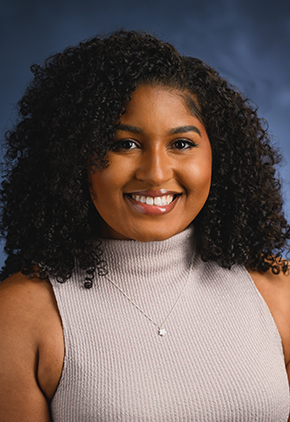 Dr. Jasmine Moore smiling for a professional headshot in front of a blue background. She has on a mauve mock turtleneck and small star necklace.