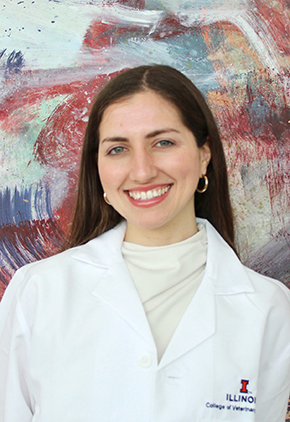 Dr. Marisa Pico smiling in front of a large abstract mural. She has on a white turtleneck and her University of Illinois whitecoat.